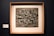 A framed artwork featuring a textured, abstract sculpture with various rectangular and geometric shapes. The piece has a three-dimensional quality with rough, uneven surfaces and a metallic appearance.