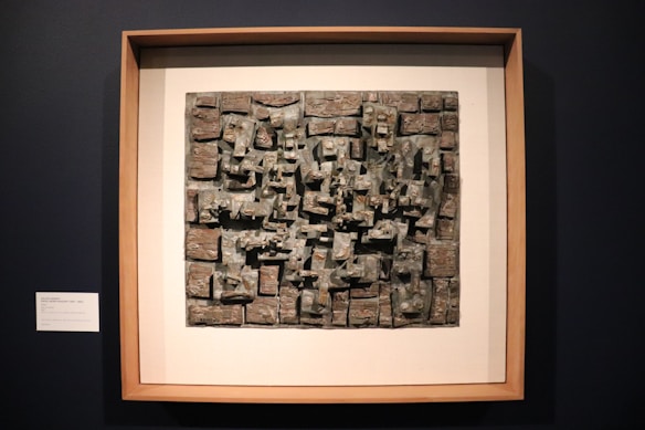 A framed artwork featuring a textured, abstract sculpture with various rectangular and geometric shapes. The piece has a three-dimensional quality with rough, uneven surfaces and a metallic appearance.