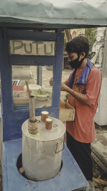 A street vendor wearing a face mask is preparing traditional cakes in a small blue stall. The vendor is working with a metal steaming pot typically used for making snacks, and a towel is draped over their shoulder. The word 'PUTU' is displayed on the stall.