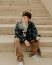 A young person with tousled hair sits on concrete steps, wearing a plaid shirt over a graphic t-shirt, brown pants, and light-colored sneakers. They appear to be holding a lit cigarette with smoke visible in the air.