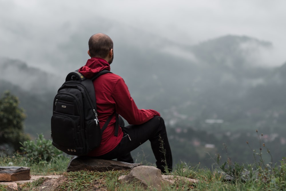 man in red jacket and black pants sitting on rock