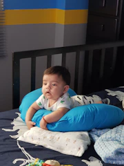A baby lies on its stomach on a bed, supported by a blue pillow. The baby is dressed in a white outfit with green patterns and has a pacifier nearby. The background shows a wall with blue, yellow, and gray stripes and a dark-colored crib. Light filters into the room from a window with blinds.