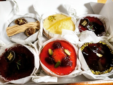 A selection of gourmet pastries, each with unique toppings and garnishes, arranged in a white box. The pastries feature a variety of textures and colors, including creamy, fruity, and nutty elements.