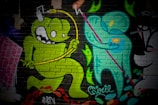 A vibrant mural on a brick wall featuring cartoon-style characters. A green monster-like figure with large eyes and a playful expression jumps energetically, holding a yellow object connected by a wire over its head. Next to it, a teal humanoid figure holding a pink staff is depicted, creating a dynamic interaction between the two characters. The background includes abstract, colorful shapes and textural elements.