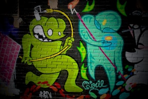 A vibrant mural on a brick wall featuring cartoon-style characters. A green monster-like figure with large eyes and a playful expression jumps energetically, holding a yellow object connected by a wire over its head. Next to it, a teal humanoid figure holding a pink staff is depicted, creating a dynamic interaction between the two characters. The background includes abstract, colorful shapes and textural elements.
