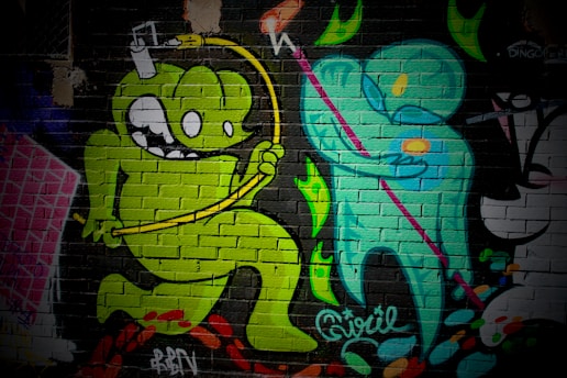 A vibrant mural on a brick wall featuring cartoon-style characters. A green monster-like figure with large eyes and a playful expression jumps energetically, holding a yellow object connected by a wire over its head. Next to it, a teal humanoid figure holding a pink staff is depicted, creating a dynamic interaction between the two characters. The background includes abstract, colorful shapes and textural elements.