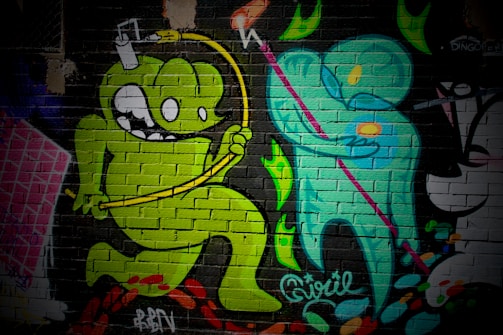 A vibrant mural on a brick wall featuring cartoon-style characters. A green monster-like figure with large eyes and a playful expression jumps energetically, holding a yellow object connected by a wire over its head. Next to it, a teal humanoid figure holding a pink staff is depicted, creating a dynamic interaction between the two characters. The background includes abstract, colorful shapes and textural elements.
