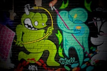 A vibrant mural on a brick wall featuring cartoon-style characters. A green monster-like figure with large eyes and a playful expression jumps energetically, holding a yellow object connected by a wire over its head. Next to it, a teal humanoid figure holding a pink staff is depicted, creating a dynamic interaction between the two characters. The background includes abstract, colorful shapes and textural elements.