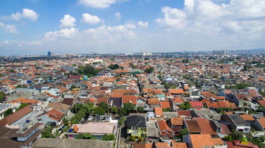 Aerial view of a densely populated urban area with numerous houses featuring red-tiled roofs. The landscape stretches into the distance, with a few taller buildings visible on the horizon. The sky is partly cloudy, adding a soft texture above the cityscape.