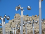 Rows of metallic anemometers are arranged against a backdrop of a rugged, eroded rock formation under a clear blue sky. The shiny silver cups are mounted on tall poles and are used for measuring wind speed.