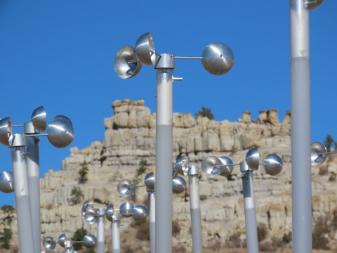 Rows of metallic anemometers are arranged against a backdrop of a rugged, eroded rock formation under a clear blue sky. The shiny silver cups are mounted on tall poles and are used for measuring wind speed.