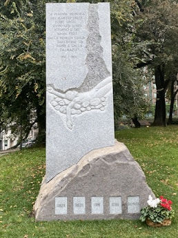 A monument made of stone with engraved text commemorating the memory of martyrs. The structure features an engraving of a rocky landscape with two hands reaching above stones. The monument is situated on a grassy area with trees in the background. A small arrangement of red and white flowers is present at the base.