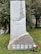 A monument made of stone with engraved text commemorating the memory of martyrs. The structure features an engraving of a rocky landscape with two hands reaching above stones. The monument is situated on a grassy area with trees in the background. A small arrangement of red and white flowers is present at the base.