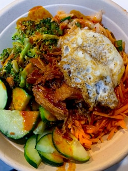 A bowl containing a variety of fresh ingredients, including sliced cucumbers, broccoli florets, shredded carrots, and pulled meat topped with a fried egg. The dish is garnished with a drizzle of sauce, adding a vibrant spice element to the colorful vegetables.