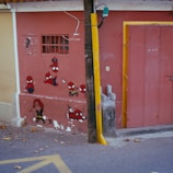 A wall is decorated with various cartoon and comic book character paintings, including multiple versions of a character with a red and blue costume, and another with a black and red costume. The scene includes a wooden utility pole in front of the wall and leaves scattered on the ground.