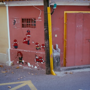 A wall is decorated with various cartoon and comic book character paintings, including multiple versions of a character with a red and blue costume, and another with a black and red costume. The scene includes a wooden utility pole in front of the wall and leaves scattered on the ground.