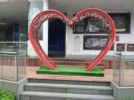 A large red heart-shaped metal structure is filled with numerous love locks, situated on a platform covered with green artificial grass. Behind the heart structure, there are signs advertising personalized love locks and a white door. The setting appears to be an urban area or a shopping center with glass railings and stairs leading up to the platform.