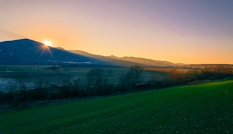 A beautiful landscape featuring a vibrant sunrise peeking over distant mountains. The foreground displays a lush green field with patches of trees and shrubs, leading to open fields. The sky transitions from a soft orange near the horizon to a clear blue, with distinct sun rays cutting through the morning mist.