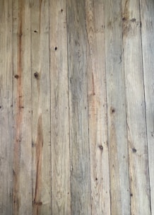 A close-up view of wooden floorboards with a natural, rustic appearance. Each plank has unique grain patterns and knots, with variations in color and texture that add character.
