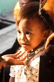 A young child with short hair is gazing intently, lit by warm sunlight. They are wearing a light-colored shirt with a pattern and appear to be interacting with an adult, as a hand is gently touching their chin. The background is out of focus, with hints of a balloon in an orange hue.