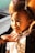 A young child with short hair is gazing intently, lit by warm sunlight. They are wearing a light-colored shirt with a pattern and appear to be interacting with an adult, as a hand is gently touching their chin. The background is out of focus, with hints of a balloon in an orange hue.