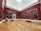 A spacious art gallery with burgundy red walls adorned with classical paintings in gold frames. The polished wooden floor reflects the soft lighting. Two people are walking through the gallery, and several benches are placed for seating.