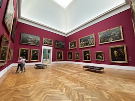 A spacious art gallery with burgundy red walls adorned with classical paintings in gold frames. The polished wooden floor reflects the soft lighting. Two people are walking through the gallery, and several benches are placed for seating.
