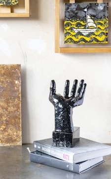 A black sculpture of an upraised hand is placed on top of two books stacked on a metallic surface. In the background, framed artwork displays a sailboat amidst stylized yellow waves against a dark sky.