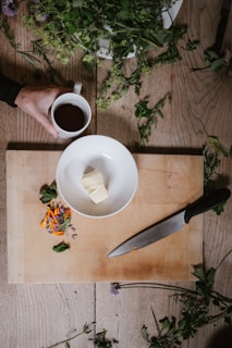 A wooden cutting board is placed on a table with a white bowl containing a few pieces of butter. Next to the bowl is a large kitchen knife with a black handle. Loose herbs and flowers are scattered around the board and on the table. A hand is holding a mug filled with a dark liquid, possibly coffee or tea.