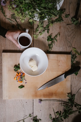 A wooden cutting board is placed on a table with a white bowl containing a few pieces of butter. Next to the bowl is a large kitchen knife with a black handle. Loose herbs and flowers are scattered around the board and on the table. A hand is holding a mug filled with a dark liquid, possibly coffee or tea.