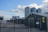 Several large metal silos are positioned next to long, green-roofed farm buildings. These structures are enclosed by a wire fence, and multiple signs are visible, indicating that the site is an approved poultry farm. The sky is partly cloudy, providing a mixture of sunlight and shade across the scene.