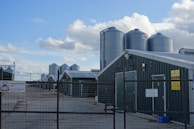 Several large metal silos are positioned next to long, green-roofed farm buildings. These structures are enclosed by a wire fence, and multiple signs are visible, indicating that the site is an approved poultry farm. The sky is partly cloudy, providing a mixture of sunlight and shade across the scene.