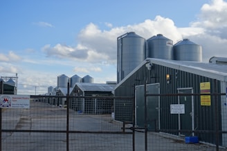 Several large metal silos are positioned next to long, green-roofed farm buildings. These structures are enclosed by a wire fence, and multiple signs are visible, indicating that the site is an approved poultry farm. The sky is partly cloudy, providing a mixture of sunlight and shade across the scene.