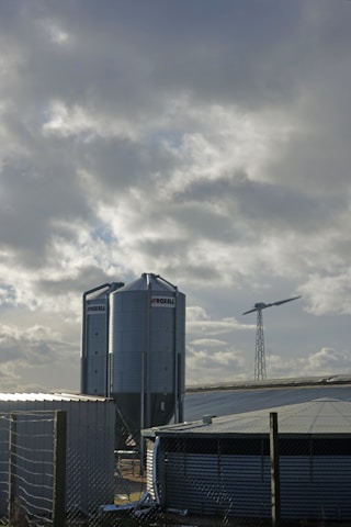 A rural landscape featuring large metallic silos with the brand name Roxell, situated near what appear to be farm buildings or structures. These silos are used for storing agricultural materials. Behind them, a single wind turbine stands tall against a cloudy sky, suggesting renewable energy use in the area.