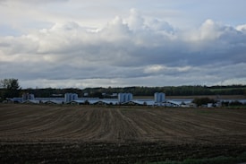 A vast agricultural landscape with freshly plowed fields stretching into the distance, bordered by rows of industrial silos and large farming structures. The sky is overcast with thick, rolling clouds, and greenery and forests can be seen in the background.