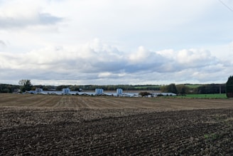 A wide expanse of farmland features a freshly plowed field in the foreground. In the background, there are several silos and farm buildings surrounded by patches of trees and expansive green pastures. The sky is overcast with large, fluffy clouds.