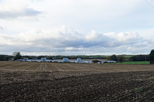 A wide expanse of farmland features a freshly plowed field in the foreground. In the background, there are several silos and farm buildings surrounded by patches of trees and expansive green pastures. The sky is overcast with large, fluffy clouds.