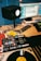 A music studio setup featuring a turntable with a vinyl record, a DJ mixer, multiple computer monitors displaying audio editing software, a studio monitor speaker, and various cables and accessories on the desk. There is also a small figurine on top of one of the monitors.