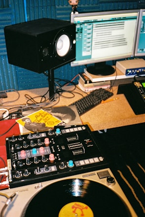 A music studio setup featuring a turntable with a vinyl record, a DJ mixer, multiple computer monitors displaying audio editing software, a studio monitor speaker, and various cables and accessories on the desk. There is also a small figurine on top of one of the monitors.