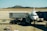 An airplane is parked at the airport receiving services from a catering truck while connected to a passenger boarding bridge. The background features a mountainous landscape under a clear blue sky.