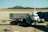 An airplane is parked at the airport receiving services from a catering truck while connected to a passenger boarding bridge. The background features a mountainous landscape under a clear blue sky.
