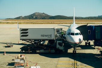 An airplane is parked at the airport receiving services from a catering truck while connected to a passenger boarding bridge. The background features a mountainous landscape under a clear blue sky.