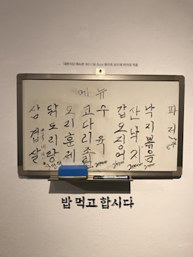 Whiteboard with handwritten Korean words arranged in rows and columns, likely representing a menu. The board also includes some prices written in smaller print at the bottom. Beneath the whiteboard, there's a phrase written on the wall in Korean. A marker and an eraser are placed on the tray attached to the bottom of the whiteboard.