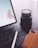 A close-up view of a workspace featuring a laptop with a stylus resting on the keyboard, a camera lens with 'Sony' branding beside it, and a smartphone with a leather case. The laptop screen displays a blurred background with app icons visible.