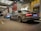 Two luxury cars are parked in a well-lit urban area with modern architecture and overhead lighting. The closer car is grey and the one further is silver, both parked side by side. The setting appears to be near a building entrance with glass doors and advertising displays.