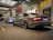 Two luxury cars are parked in a well-lit urban area with modern architecture and overhead lighting. The closer car is grey and the one further is silver, both parked side by side. The setting appears to be near a building entrance with glass doors and advertising displays.