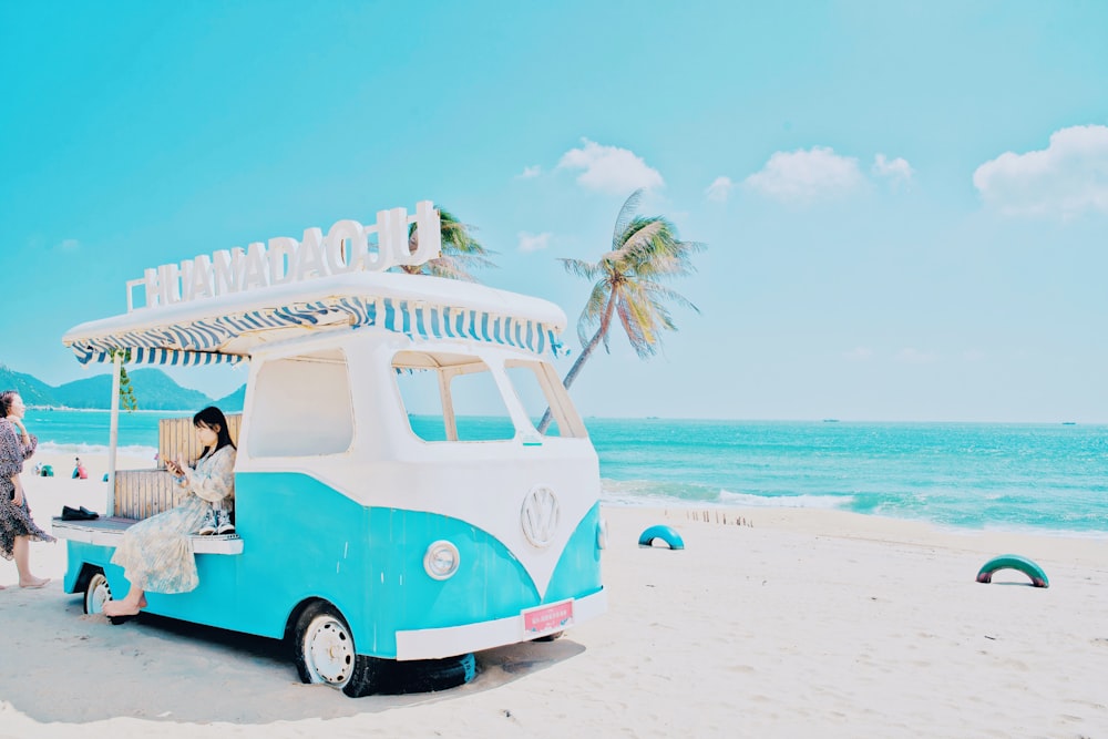 man in white t-shirt sitting on white and blue volkswagen t-1 on beach during