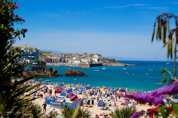 A bustling beach scene with numerous people sunbathing, swimming, and enjoying the sunny weather. Vibrant beach umbrellas and tents dot the sandy shore, while the clear blue sea supports several small boats. A picturesque coastal town with white buildings is visible in the background, bordered by rocky outcrops.