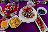 A beautifully arranged table setting features a chocolate cake drizzled with white chocolate and topped with fresh strawberries and raspberries. Surrounding items include a bowl of blueberries, a platter of strawberries, a vase with red roses and other flowers, and savory snacks like nachos. Also present are apples, a bottle of wine, and a pot of tea, all set on a rich purple tablecloth.
