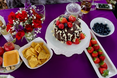 A beautifully arranged table setting features a chocolate cake drizzled with white chocolate and topped with fresh strawberries and raspberries. Surrounding items include a bowl of blueberries, a platter of strawberries, a vase with red roses and other flowers, and savory snacks like nachos. Also present are apples, a bottle of wine, and a pot of tea, all set on a rich purple tablecloth.